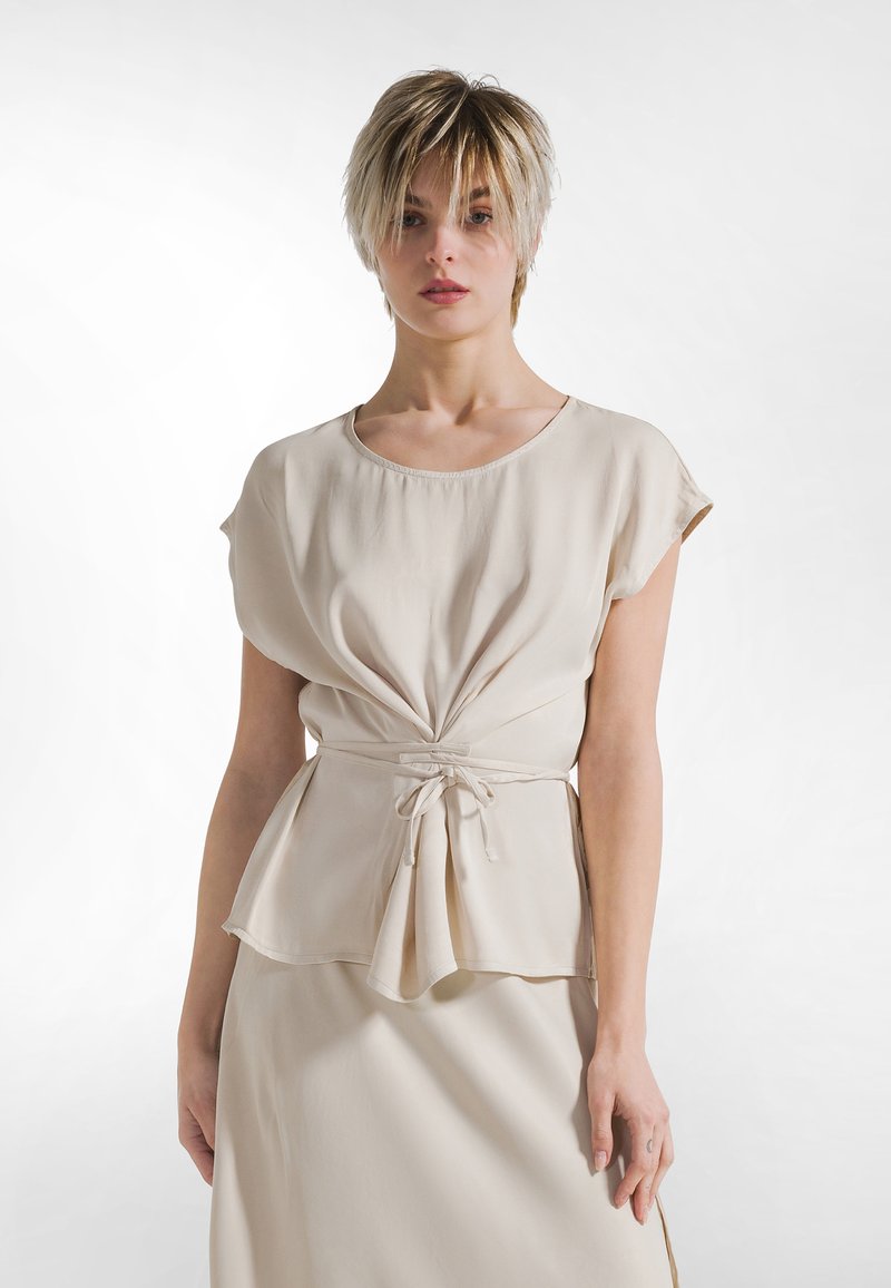 Beige, let top med korte ærmer. Har en rynket front og et bånd ved taljen. Glat tekstur med en afslappet pasform.