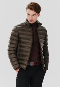 Ciemnobrązowa kurtka puchowa z wysokim kołnierzem, poziomym pikowaniem, zapięciem na zamek oraz dopasowanym krojem. Nosi się ją na ciemnym golfie.