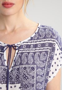 Bílá blůza s fialovými složitými paisley a květinovými vzory, krátké rukávy, výstřih ve tvaru klíčové dírky se zavazováním a jemným textilním povrchem.