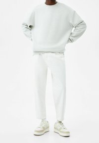 Sudadera gris claro con cuello redondo, combinada con pantalones blancos. El calzado presenta acentos en beige y blanco. Diseño minimalista, tela texturizada.