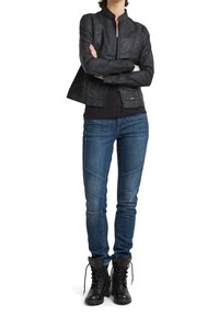 Kvinna med armarna i kors, klädd i en svart jacka, mörk skjorta, åtsittande blå jeans med sömdetaljer och svarta snörstövlar.