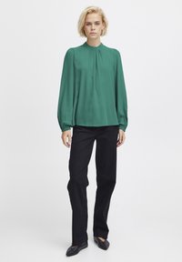 Blusa verde a maniche lunghe con colletto alto, dettagli arricciati al collo, abbinata a pantaloni neri a gamba dritta e ballerine nere.