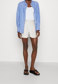 Camisa de manga larga a rayas azul claro y blanca, camiseta sin mangas blanca, pantalones cortos crema con finas rayas verticales y sandalias planas negras.