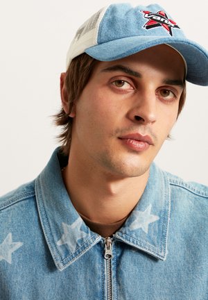 Giovane con occhi marroni che indossa una giacca di jeans blu con motivo a stelle e un berretto a rete blu e bianco con un logo rosso e nero.