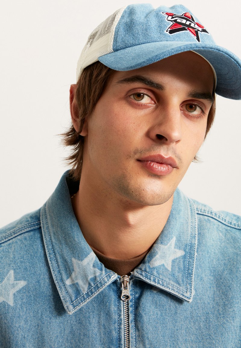 Junger Mann mit braunen Augen, trägt eine blaue Jeansjacke mit Sternenmuster und eine blau-weiße Netzkappe mit einem rot-schwarzen Logo.