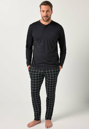Chemise noire à manches longues avec un col henley, associée à un pantalon de pyjama ajusté à motif vichy dans des tons sombres, pieds nus visibles.