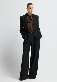 Zwarte op maat gemaakte pak met brede broekspijpen, voorzien van een bijpassende blazer en een bruine blouse met een striknek. Soepele textuur en streepjesmotief.