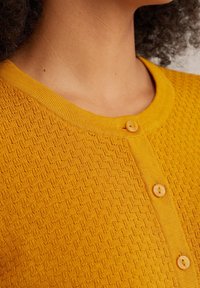 Gele gebreide cardigan met een ronde hals, voorzien van een sluiting met knopen gemaakt van hetzelfde gele materiaal en een subtiel patroon over het geheel.