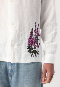 White linen shirt featuring pink floral embroidery on the lower left side, with a classic collar and button-down front.