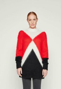 Femme portant un pull à manches longues en peluche avec des motifs géométriques rouges, blancs et noirs, se tenant devant un fond uni clair.