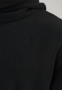 Sweatshirt en polaire noire avec col montant, présentant un logo en relief subtil au centre. Le matériau possède une surface douce et texturée.