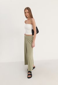 Witte strapless top, lichtgroene geplooide wijde broek en zwarte sandalen met gespen. Model houdt een kleine zwarte crossbody tas vast.