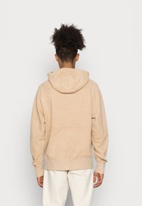 Beige cotton hoodie with a drawstring hood, ribbed cuffs, and hem. Features a subtle speckled texture and a logo tag on the neck seam.