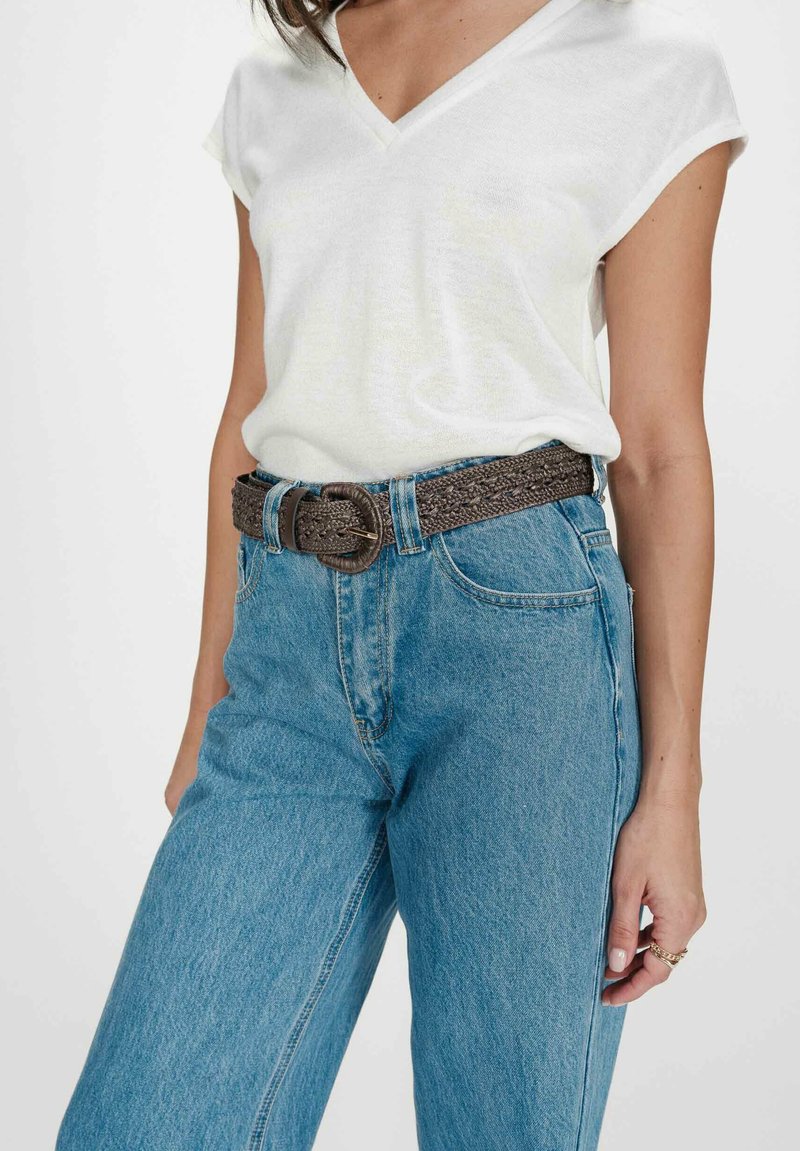 Femme portant un T-shirt blanc à manches courtes et encolure en V, rentré dans un jean taille haute bleu avec une ceinture tressée foncée.