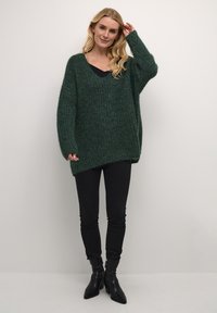Tmavě zelený oversized svetr s výstřihem do V, texturovaným pleteným materiálem a dlouhými rukávy, kombinovaný s úzkými černými kalhotami a kotníkovými botami.
