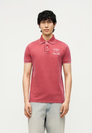 Junger Mann mit lockigem dunklem Haar, der ein ausgewaschenes rotes Hollister Malibu Polohemd und hellblaue Jeans trägt und vor einem schlichten Hintergrund steht.