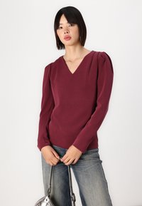 Burgunderfarget langermet topp med V-hals og pufferte skuldre, kombinert med blå jeans og en sølvveske, vist mot en nøytral bakgrunn.