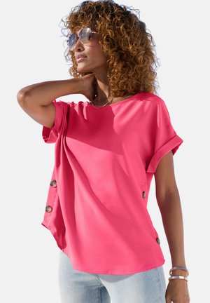 Frau mit lockigem Haar, die eine Sonnenbrille trägt und eine leuchtend pinke, kurzärmelige Bluse mit Knöpfen an der Seite sowie hellblaue Jeans anhat.