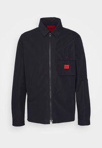 Veste noire zippée avec col, une grande poche à rabat sur la poitrine comportant un patch logo rouge Hugo, et une poche zippée sur la manche gauche.