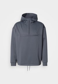 Siv hoodie z višjim ovratnikom, polovičnim zadrgo, sprednjim žepom in vrvico na dnu. Značilna je subtilna oznaka na zgornjem levem prsnem delu.
