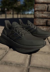 Чифт черни маратонки Adidas, поставени на открит тротоар близо до тухлена стена под естествена светлина.