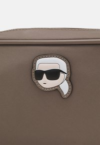 Borsa in nylon marrone con una toppa di un cartone animato che raffigura un personaggio con occhiali da sole e capelli bianchi, bordata con cuciture beige per un tocco di dettaglio in più.