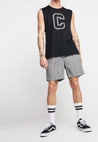T-shirt sans manches noire avec la lettre blanche "C", short à carreaux gris, baskets noires et chaussettes blanches avec des rayures noires. Tenue décontractée.