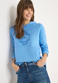 Lichtblauwe hoodie met een voorkantgrafiek in donkerder blauw. Lange mouwen, een capuchon met trekkoord en een relaxte pasvorm, gecombineerd met blauwe spijkerbroek.
