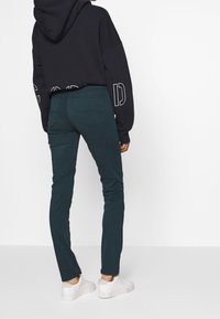 Dunkelgrüne, enganliegende Hose aus glattem Stoff mit Gesäßtaschen. Kombiniert mit einem lockeren schwarzen Kapuzenpullover und weißen Sneakern.