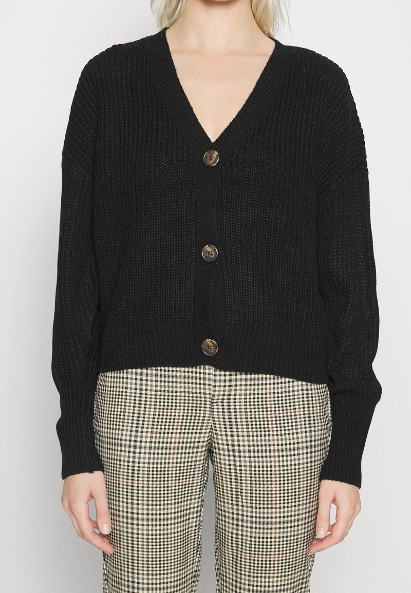 Černý pletený cardigan s výstřihem do V, třemi velkými hnědými knoflíky a dlouhými rukávy. Nosený přes kostkované kalhoty s texturovaným vzorem.