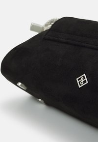 Czarna zamszowa kopertówka z srebrnym logo. Posiada zamek błyskawiczny i gładką teksturę, co podkreśla minimalistyczny design.