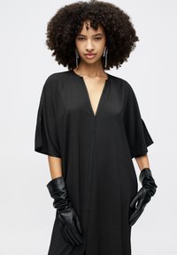 Crna kaftan haljina s dubokim V-izrezom, širokim rukavima i laganom tkaninom. Model nosi crne kožne rukavice kao istaknutu dodatak.