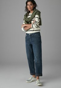 Top léger à rayures, manches longues, en blanc et bleu marine, associé à un jean en denim foncé et des baskets grises, rehaussé par un pull en maille vert.