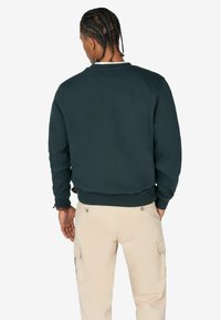 Donkergroene sweatshirt met een ronde hals, ribbels aan de manchetten en onderkant. De stof lijkt zacht en textuur te hebben, gedragen met lichtbeige broeken.