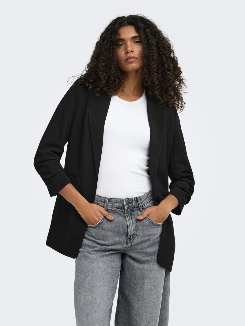 Femme aux cheveux bouclés portant un blazer noir, une chemise blanche et un jean gris, les mains dans les poches, sur un fond uni.