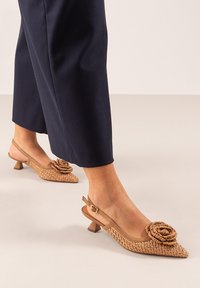Tacones slingback en tono beige tejido, con un acento floral, diseño de punta puntiaguda y un tacón bajo. Textura suave con un delicado detalle de rosa.