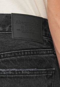 Černé džíny z denimu mají koženou nášivku na pasu s raženým textem. Podrobnosti švů jsou viditelné podél okrajů a kapes.