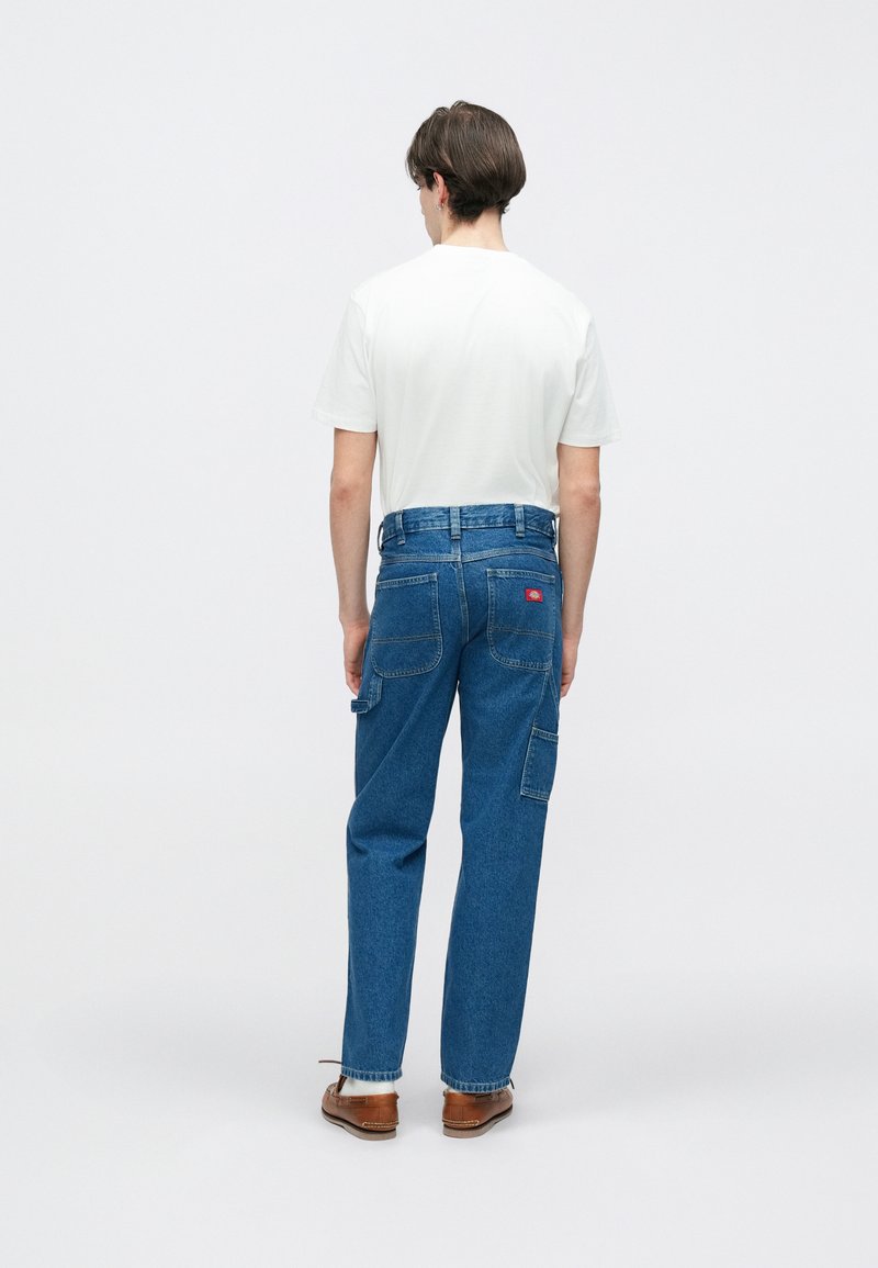 Pantalon cargo en denim bleu à coupe ample, avec des poches arrière, une ceinture et un léger évasement aux poignets. Porté avec un t-shirt blanc.