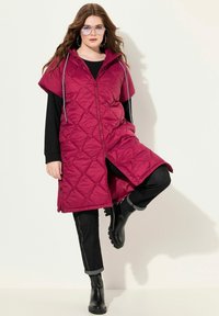 Žena v dlouhé, prošívané, burgundské bezrukávové bundě přes černé oblečení, černé boty a velké kruhové náušnice, pózující s jednou nohou zvednutou.