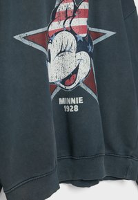 Donkerblauwe sweatshirt met een afbeelding van een glimlachend cartoonfiguurtje tegen een achtergrond van rode, witte en blauwe sterren en de tekst "MINNIE 1928."