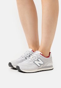 Nohy ve světlých šedých teniskách New Balance s bílými tkaničkami, černými podrážkami a červeným vnitřním lemem, stojící na bílé ploše.