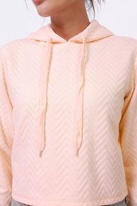 Sweatshirt court à capuche en pêche clair, avec une texture en chevron, des cordons avec embouts en métal, et une finition en tissu doux et lisse.