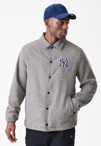 Άνδρας που φοράει σακάκι με καρό σχέδιο houndstooth και λογότυπο της New York Yankees, με μπλε καπέλο, κοιτάζοντας στο πλάι με το ένα χέρι στην τσέπη.