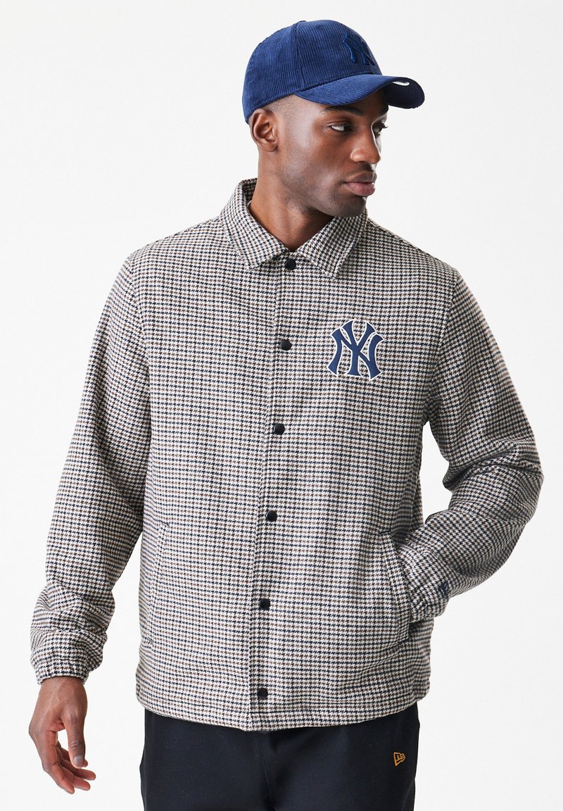 Άνδρας που φοράει σακάκι με καρό σχέδιο houndstooth και λογότυπο της New York Yankees, με μπλε καπέλο, κοιτάζοντας στο πλάι με το ένα χέρι στην τσέπη.