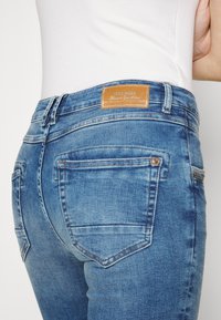 Närbild av en person som bär ljusblå denimjeans med bakfickor och en brun läderlogotyp, tillsammans med en vit topp.