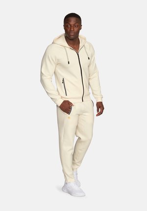 Mężczyzna ubrany w kremową bluzę na zamek i dopasowane spodnie dresowe oraz białe tenisówki, stoi z jedną ręką w kieszeni.