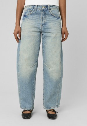 Personne portant un jean bleu clair à jambes larges avec des détails légèrement délavés et des ballerines noires, debout devant un fond gris uni.