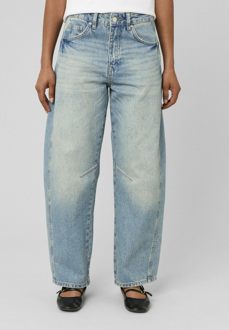 Personne portant un jean bleu clair à jambes larges avec des détails légèrement délavés et des ballerines noires, debout devant un fond gris uni.