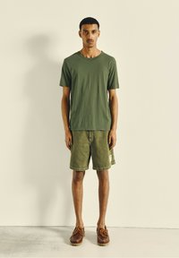 Green cotton t-shirt paired with olive shorts, both relaxed fit. Brown shoes with lace detailing; minimalistic design, smooth textures.
