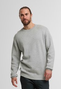 Uomo barbuto con i capelli legati dietro, che indossa un maglione a maniche lunghe grigio chiaro e pantaloni scuri, in piedi davanti a uno sfondo bianco uniforme.
